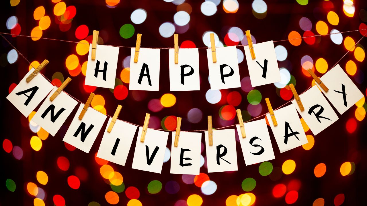 The words "happy anniversary wishes for husband" displayed on white cards hanging from a string with clothespins against a blurred background of multicolored lights.